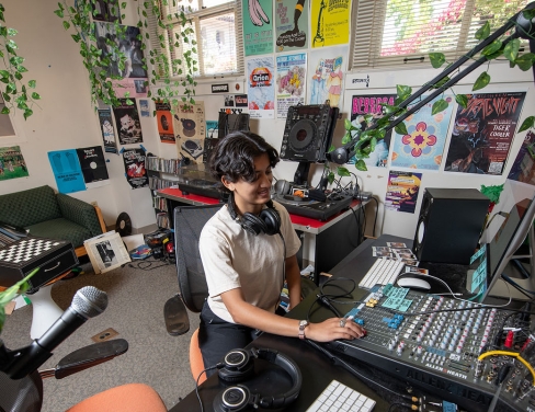 An Occidental College student in the KOXY radio station, at the DJ booth with plants all around