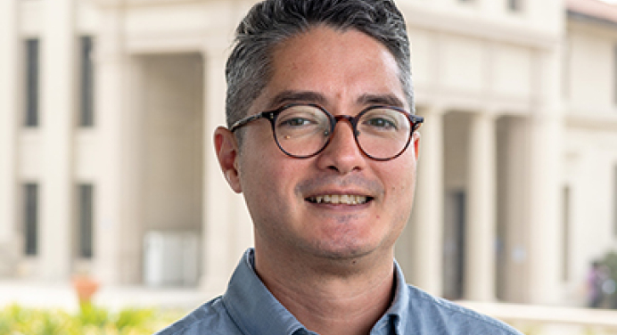 Joel is wearing a blue shirt and is standing in front of a building at the Occidental College campus