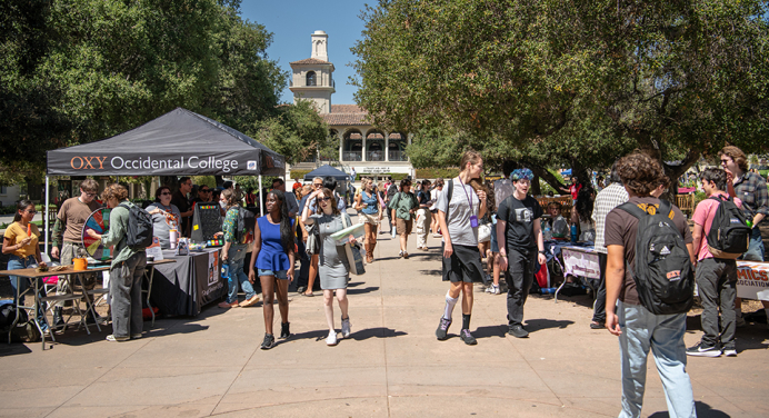  A bustling outdoor Involvement Fair at Occidental College. Students walk through a sunny campus plaza lined with club recruitment tables and an 'OXY' branded tent, showcasing a vibrant and active student life.