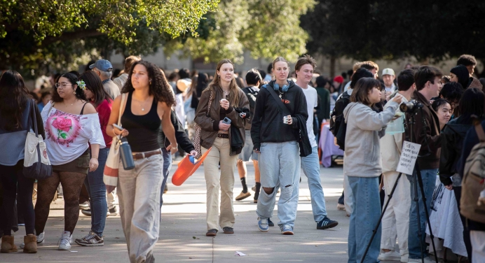 A bustling outdoor campus walkway filled with students, some walking and others stopping at tables for conversations. Trees with green foliage provide shade along the path. Students are casually dressed, carrying backpacks, water bottles, and tote bags.