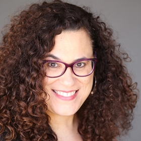 Rachel has brown curly hair and is wearing dark red glasses.