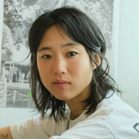 Joanne is wearing a white top, has dark hair and is turned, looking at the camera