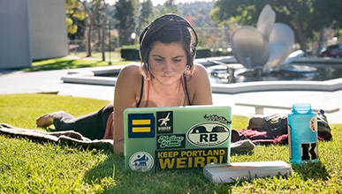 A student with headphones looks at her laptop while lying on the quad in front of Gilman Fountain