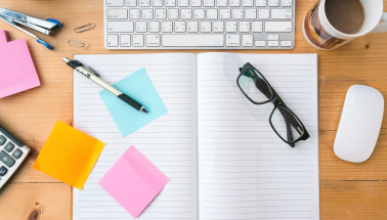 top-down view of an organized wooden office desk with various items including a keyboard, mug, notebook and colorful sticky notes