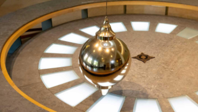 A Foucault pendulum: a large, shiny, brass-colored bob (ball) suspended by a wire over a circular floor display with light-up segments and a star design at the center