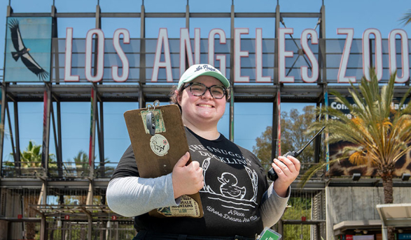Oxy student intern with a clipboard in front of the Los Angeles Zoo sign