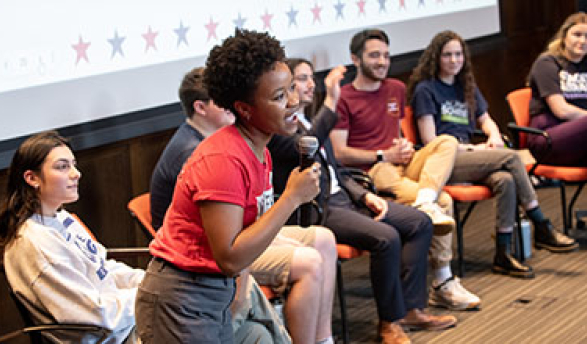 a student stands with a mic in front of a panel of other students