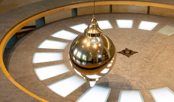 A Foucault pendulum: a large, shiny, brass-colored bob (ball) suspended by a wire over a circular floor display with light-up segments and a star design at the center