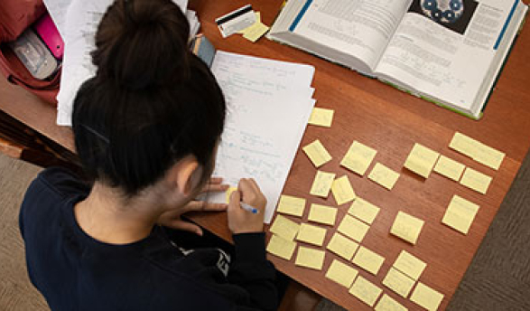 A student studies for final exams at a desk covered with papers and sticky notes