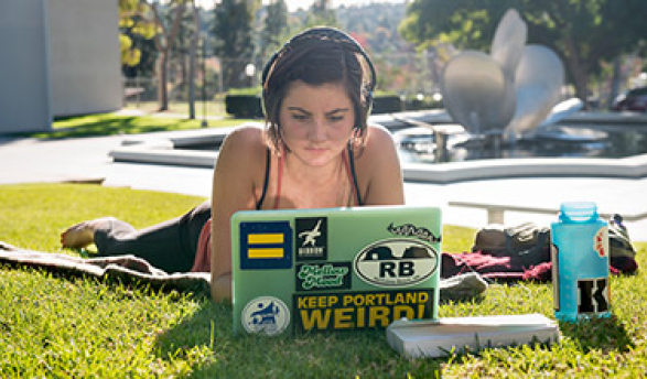 A student with headphones looks at her laptop while lying on the quad in front of Gilman Fountain