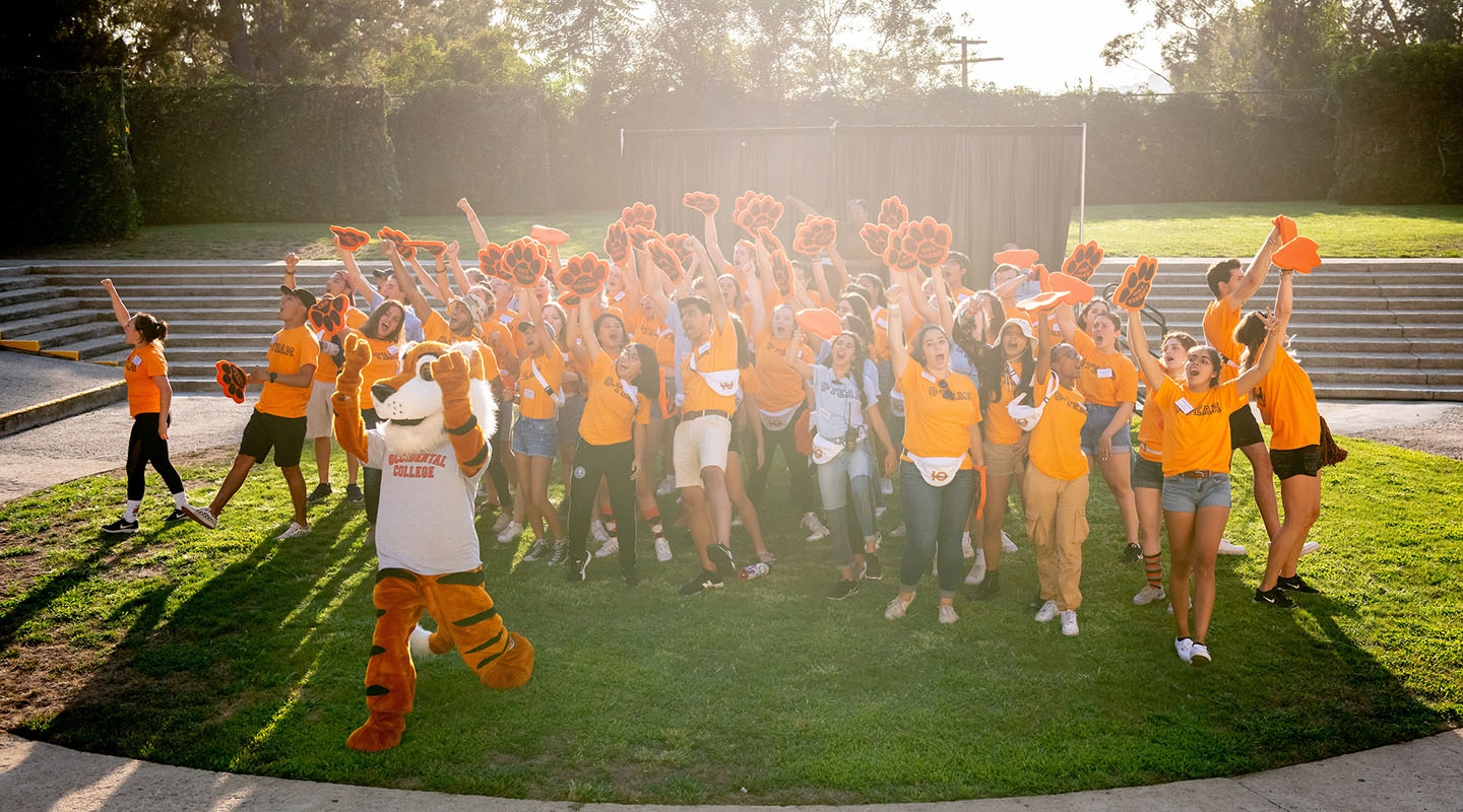 A crowd of Occidental College O-Team orientation leaders, cheering in a green field with Oswald in front