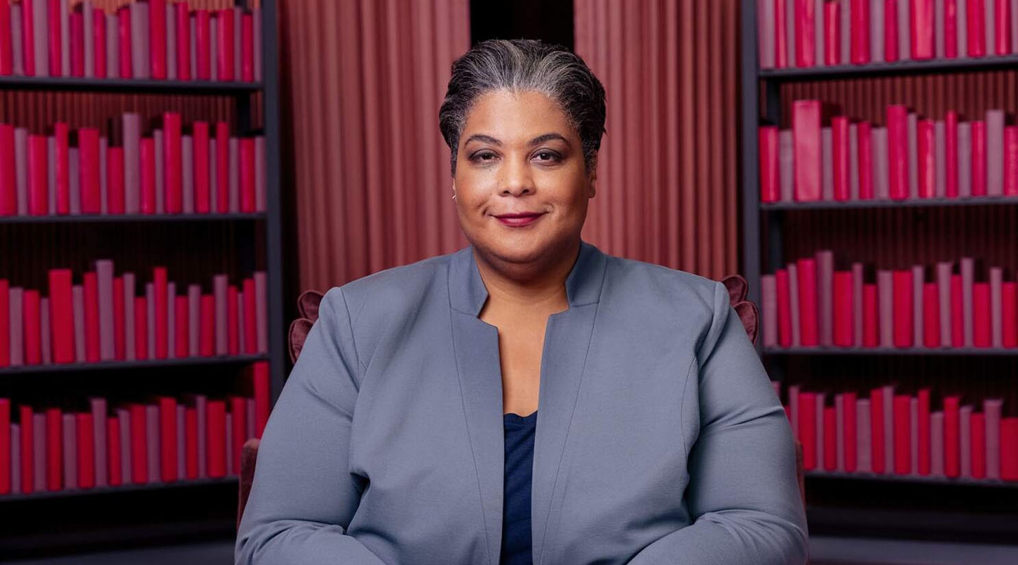 Roxane Gay headshot in a grey suit against a burgundy background