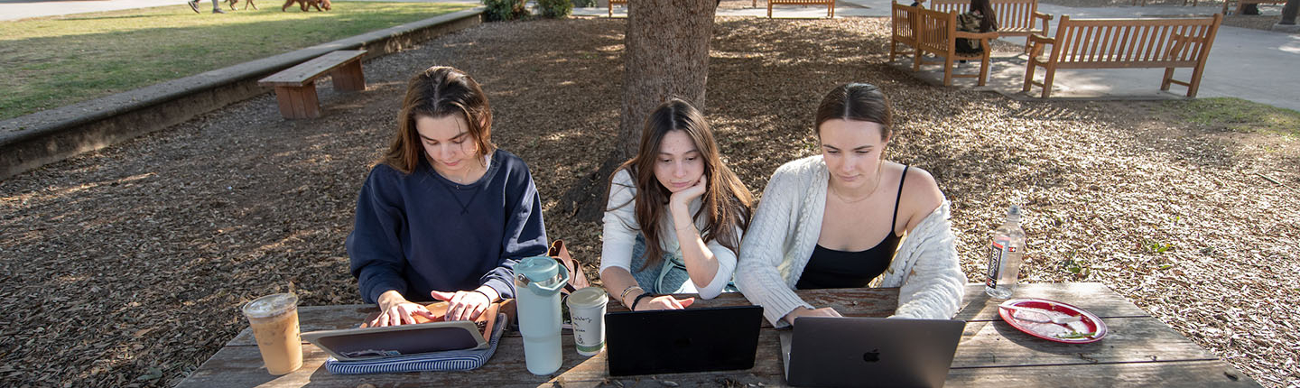 Three Occidental College students sitting side by side at a picnic table, looking at their laptops