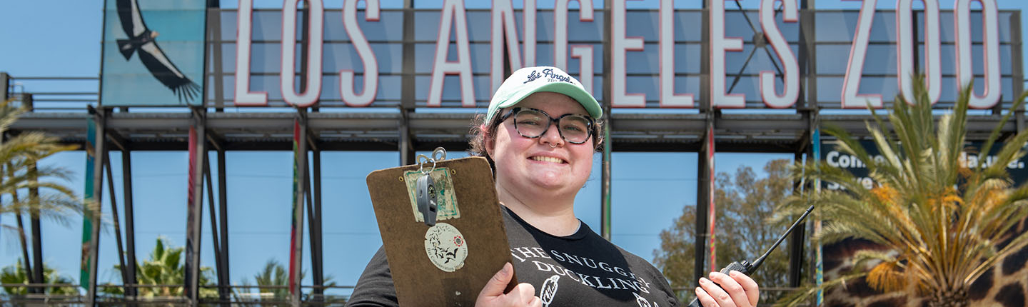 Occidental College student intern standing in front of the Los Angeles Zoo sign with a clipboard and hat