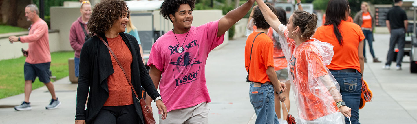An O-Team member hi-fives a new first-year student walking on the sidewalk with his mother
