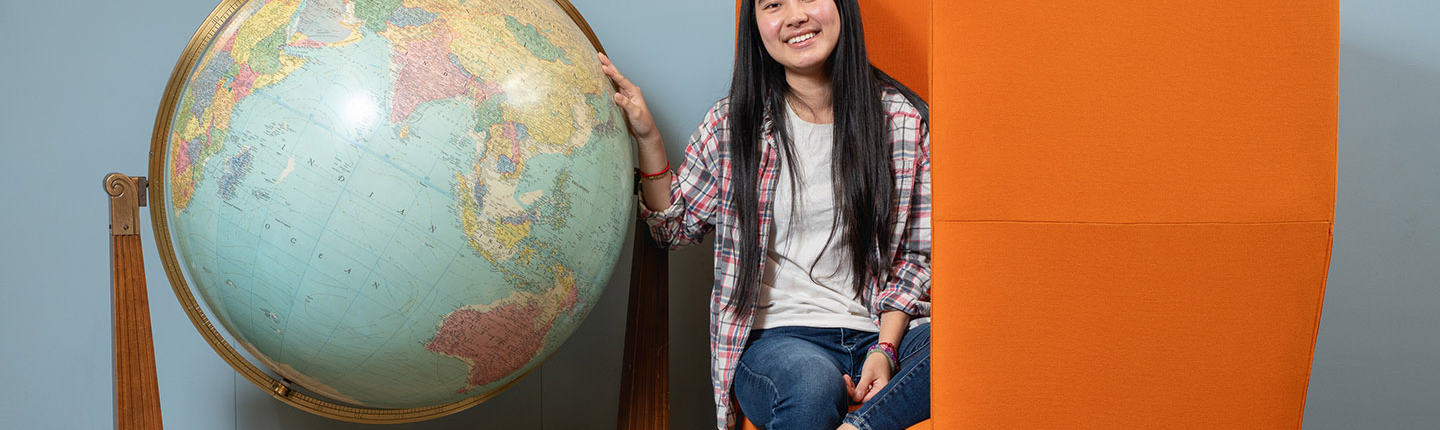 Thina Ly '27 sits next to a giant globe in the campus library, seated on an orange chair