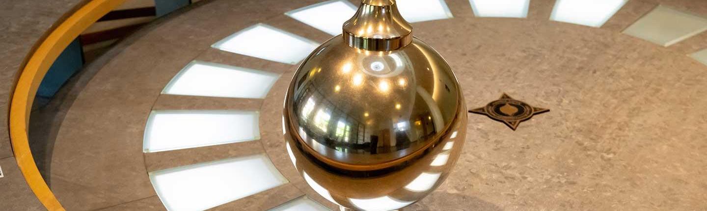 A Foucault pendulum: a large, shiny, brass-colored bob (ball) suspended by a wire over a circular floor display with light-up segments and a star design at the center