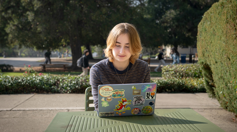 An Occidental College student sits at an outdoor table looking at her laptop.