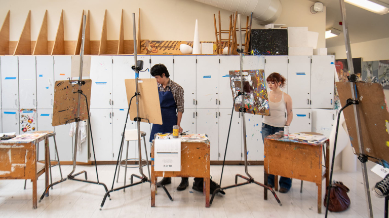 Two Occidental students painting at their easels standing in the art studio
