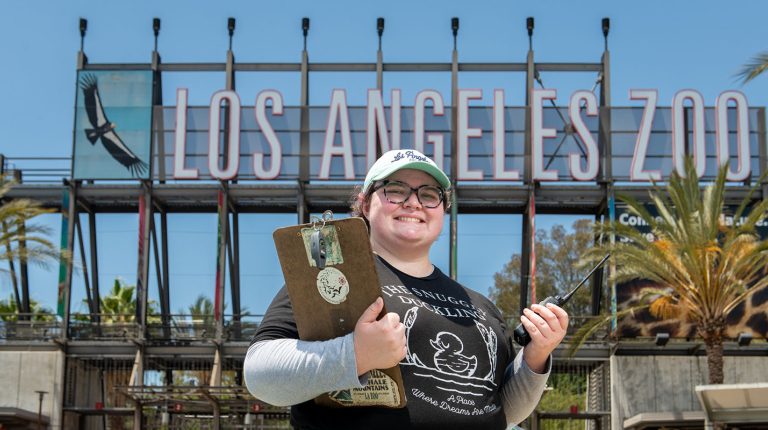 Occidental College student intern standing in front of the Los Angeles Zoo sign with a clipboard and hat