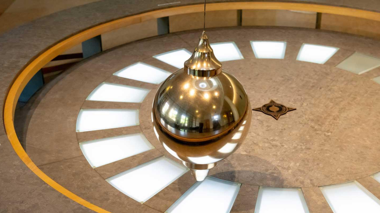 A Foucault pendulum: a large, shiny, brass-colored bob (ball) suspended by a wire over a circular floor display with light-up segments and a star design at the center