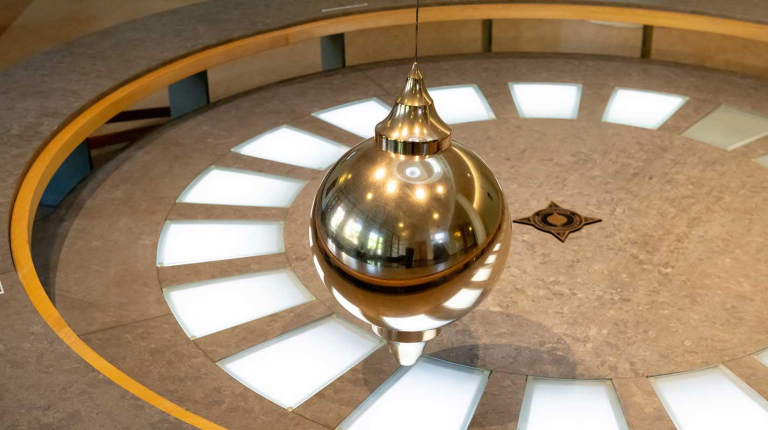 A Foucault pendulum: a large, shiny, brass-colored bob (ball) suspended by a wire over a circular floor display with light-up segments and a star design at the center