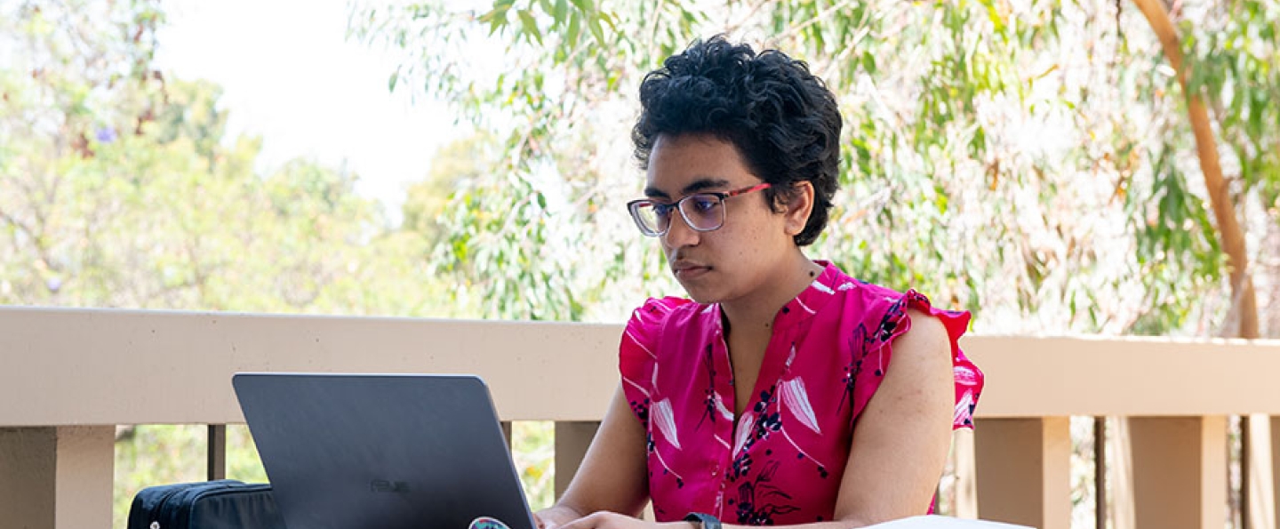  A student looks intently at a laptop in the foreground, with lush foliage in the background