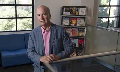 Derek Shearer in a blue blazer in front of a bookshelf and a window on campus