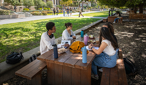 Occidental (Oxy) students sitting at wood table on a green campus with buildings in the background