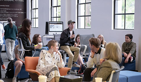 A group of Occidental (Oxy) students and a professor seated around a room in discussion. One person holds a microphone.