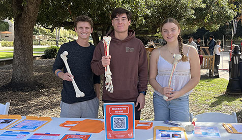Three Occidental College kinesiology students at a table holding plastic models of bones