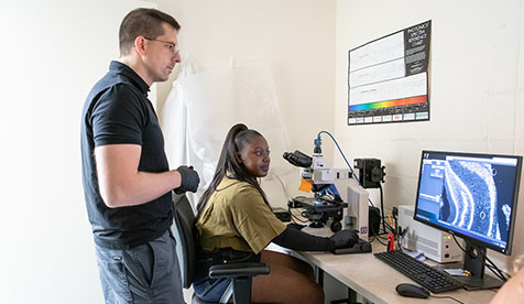 an Occidental (Oxy) student is seated at a microscope with a professor standing nearby