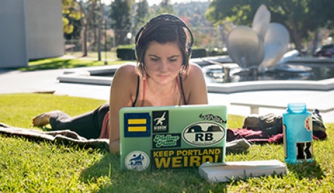 A student with headphones looks at her laptop while lying on the quad in front of Gilman Fountain