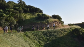 A wide image photograph of a group of people walking along a dirt path on a green hillside. 