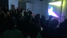 A group of people is seated watching a screening in the OXY ARTS gallery. The room is dark and the screen is lit with bright abstract colors. 