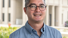 Joel is wearing a blue shirt and is standing in front of a building at the Occidental College campus