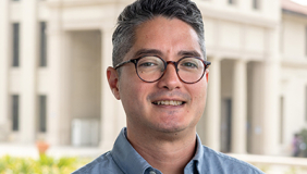 Joel is wearing a blue shirt and is standing in front of a building at the Occidental College campus
