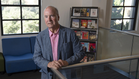 Derek Shearer in a blue blazer in front of a bookshelf and a window on campus