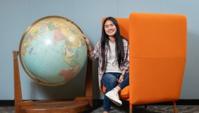 Thina Ly '27 sits next to a giant globe in the campus library, seated on an orange chair