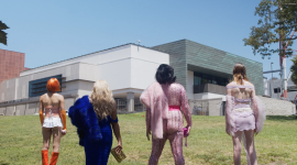 A still image of performers in vibrant, avant-garde outfits standing in front of a building. 