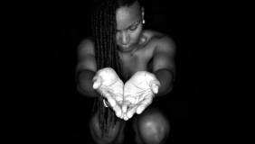 A black-and-white portrait showing a figure kneeling in darkness with long braided hair, gazing down at their own cupped hands extended toward the viewer and illuminated by a single light source against an otherwise black background.