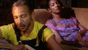 A man wearing overalls and a yellow shirt is in the foreground looking down. Behind him, a woman lies on a brown couch, wearing a purple dress, eyes shut.
