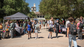  A bustling outdoor Involvement Fair at Occidental College. Students walk through a sunny campus plaza lined with club recruitment tables and an 'OXY' branded tent, showcasing a vibrant and active student life.