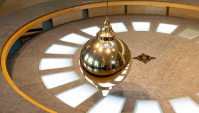 A Foucault pendulum: a large, shiny, brass-colored bob (ball) suspended by a wire over a circular floor display with light-up segments and a star design at the center