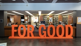 Large wooden orange sign reading "FOR GOOD" rests in front of a library circulation desk. 