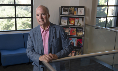 Derek Shearer in a blue blazer in front of a bookshelf and a window on campus
