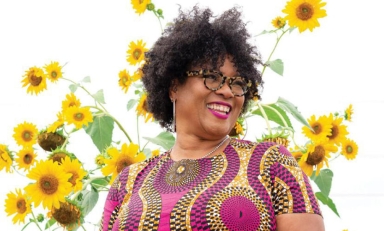 Monica White headshot in a colorful dress in front of tall sunflower plants