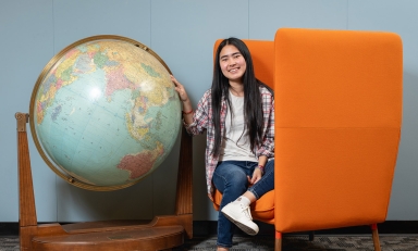 Thina Ly '27 sits next to a giant globe in the campus library, seated on an orange chair