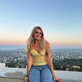 Grace is sitting on a wall with the city and sunsetting sky in background