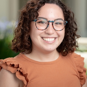 Image displays a woman with shoulder-length brunette curly hair and glasses wearing an orange shirt